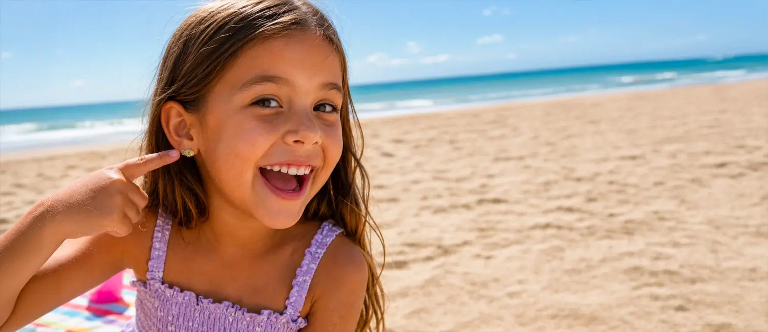 A young girl with long brown hair smiles pointing to her Kaykies fish earring on a sunny beach, wearing a light purple ruffled spaghetti-strap top. The background shows a sandy beach and the blue ocean under a clear sky.