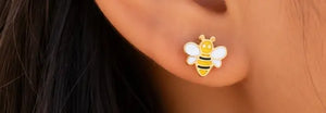 A close-up of a person's ear wearing a small bee stud earring. The earring is yellow and black striped with white wings and a gold-tone outline.