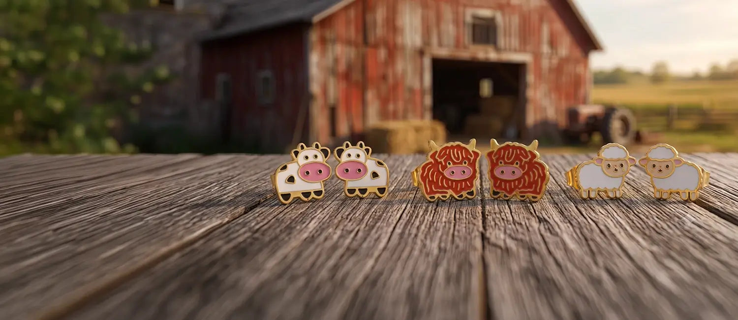 Four small animal-shaped enamel earrings, including four cows and two sheep, arranged on a weathered wooden surface with a blurred red barn and outdoor field in the background.