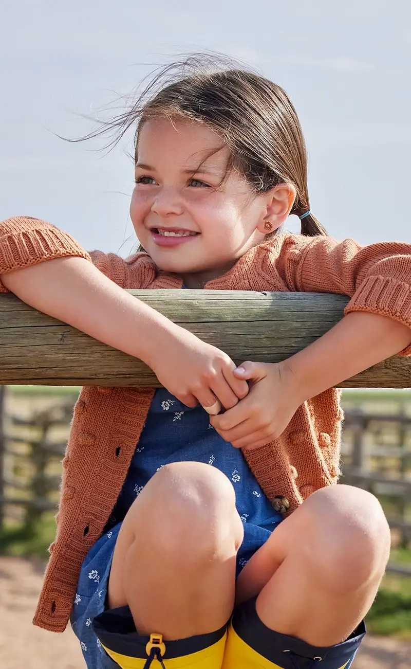 A young girl with brown hair in a ponytail smiles while leaning on a wooden fence outdoors wearing her cow earrings from Kaykies. She is wearing a rust-orange knit cardigan, a blue patterned dress, and yellow rain boots.